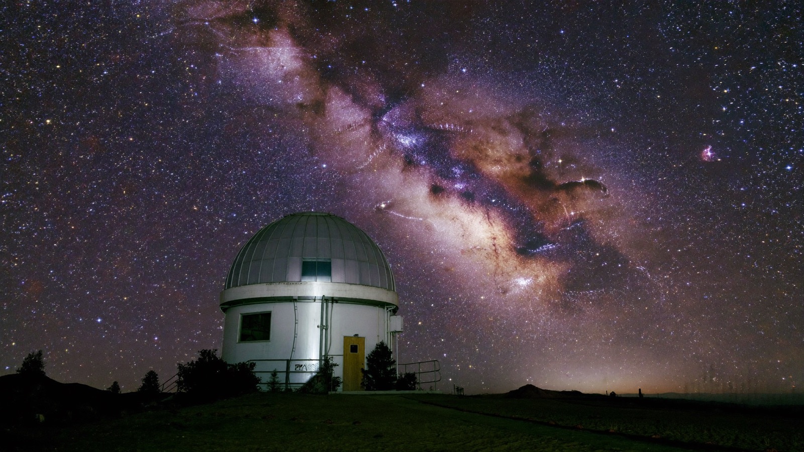 The Milky Way arching over the small observatory dome in the dark meadow behind the planetarium
